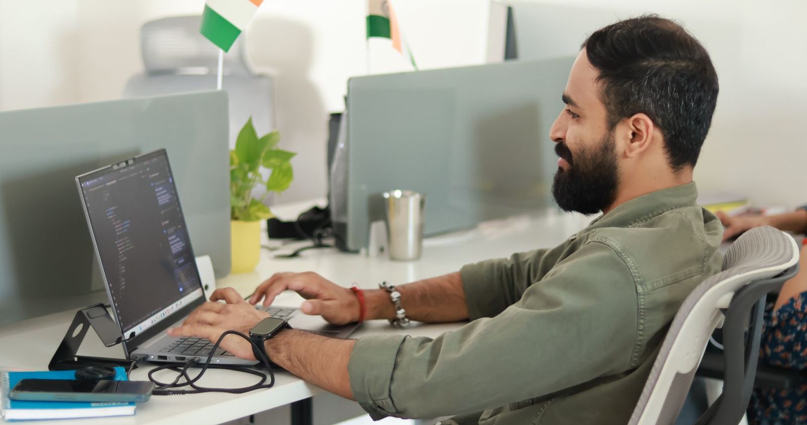 An Indian software developer coding on a laptop at his desk in an Indian office, with Indian flags on the workstation